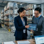Supply chain managers analyzing returns logistics administrative bottlenecks on a tablet in a modern warehouse.