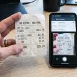 Eine Hand hält einen Beleg neben einer mobilen App für Spesenabrechnungen in einer modernen Büroumgebung – Best Practices der Verarbeitung.