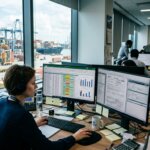 Logistics employee analyzing data for short-term ocean freight deals in a busy office overlooking a container terminal.